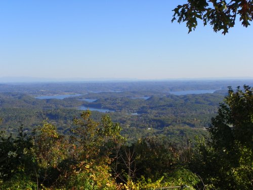 A panoramic view of rolling hills and lakes under a clear blue sky, framed by trees in the foreground.