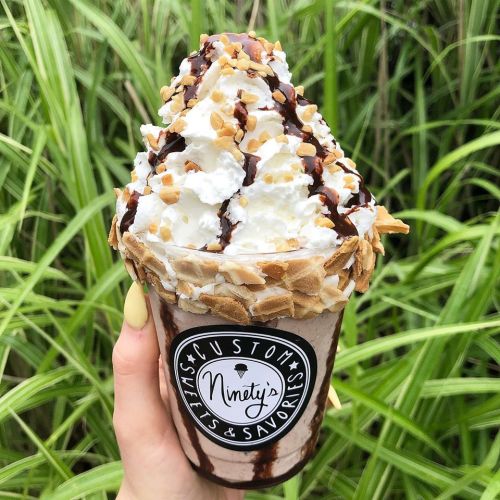 A hand holding a tall dessert cup filled with whipped cream, chocolate drizzle, and crushed nuts, set against green foliage.