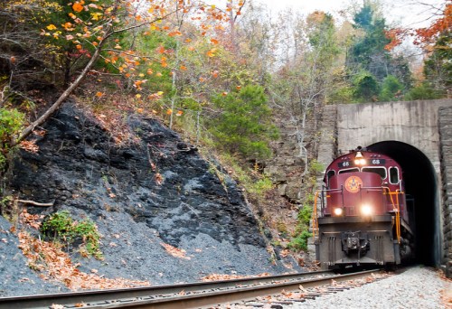 A train emerges from a tunnel surrounded by autumn foliage and rocky terrain.