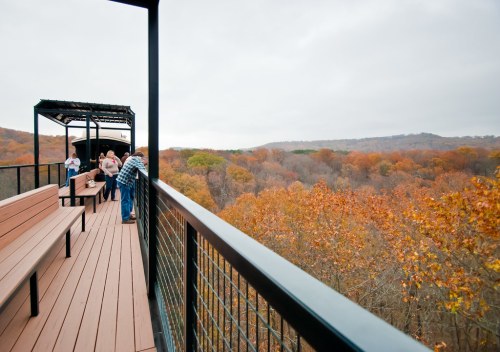 A viewing platform with people overlooking a forest of autumn-colored trees under a cloudy sky.