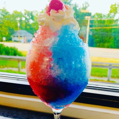 A colorful snow cone in a glass, featuring red and blue layers topped with whipped cream and a cherry.