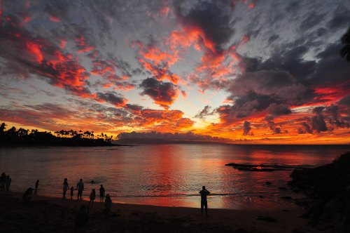 A vibrant sunset over the ocean, with colorful clouds and silhouettes of people on the beach.