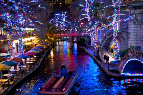 A vibrant river scene at night, adorned with colorful lights on trees and buildings, with a boat gliding through the water.