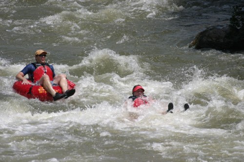 Two people enjoying a river adventure, one in a red inflatable tube and the other floating in the water, surrounded by waves.