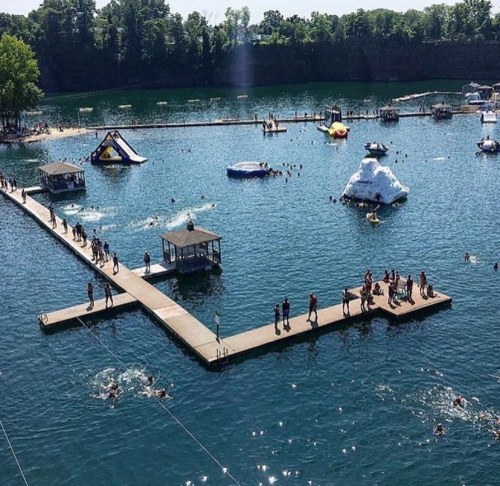 A busy lake with people swimming, a floating obstacle course, and a dock surrounded by trees on a sunny day.