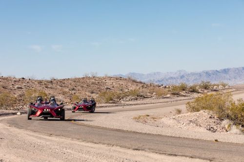 Two red three-wheeled vehicles drive along a winding desert road with mountains in the background.