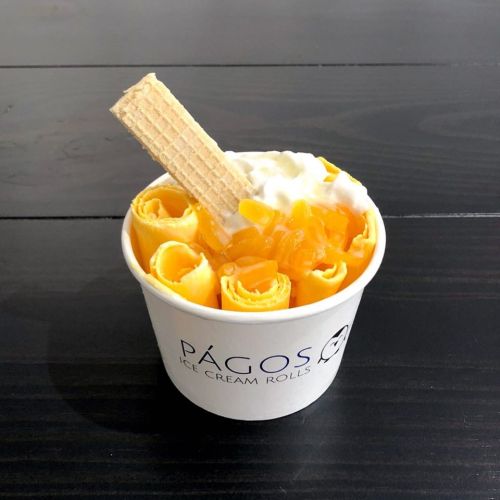 A cup of rolled ice cream topped with fruit, whipped cream, and a wafer stick on a dark wooden surface.