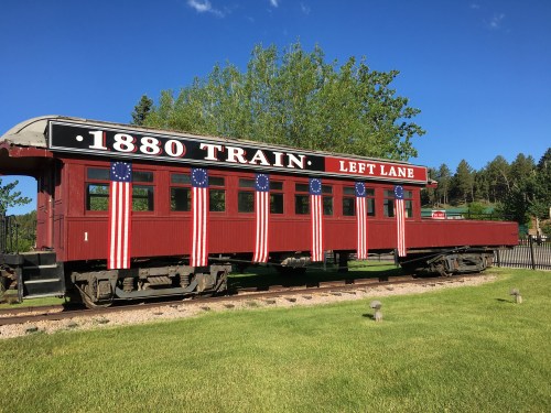 A vintage red train car decorated with American flags, labeled "1880 TRAIN LEFT LANE," set against a clear blue sky.
