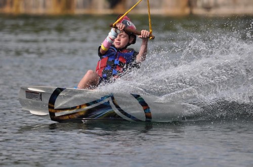 A child wearing a helmet and life jacket is wakeboarding, splashing water as they glide across a lake.