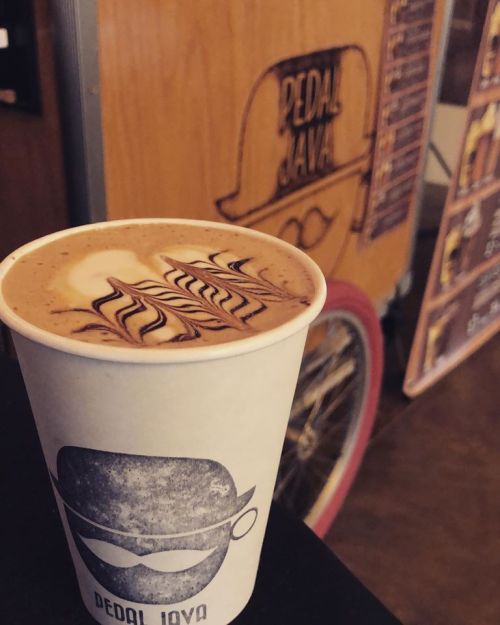 A coffee cup with latte art sits on a table, with a bicycle and a sign in the background.
