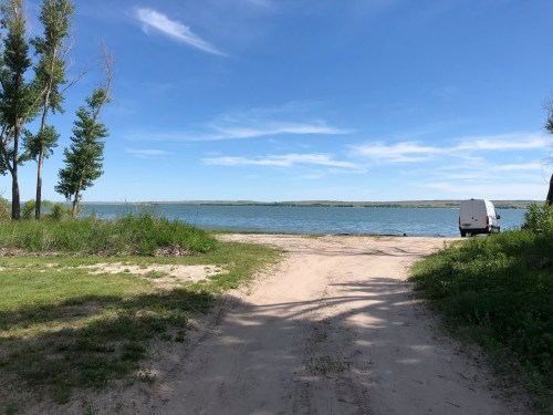 A sandy path leads to a calm lake, with trees on the left and a white van parked nearby under a clear blue sky.