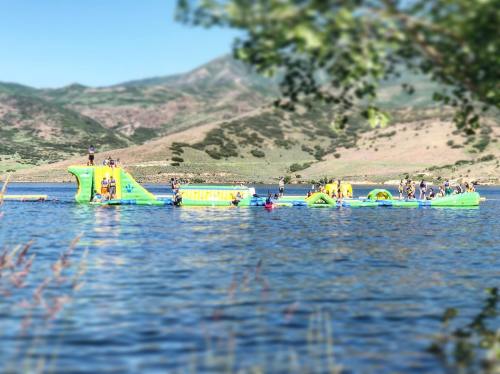 A colorful inflatable water park with people playing on it, set against a scenic lake and mountains in the background.