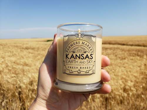 A hand holds a Kansas-themed candle in a wheat field under a clear blue sky.