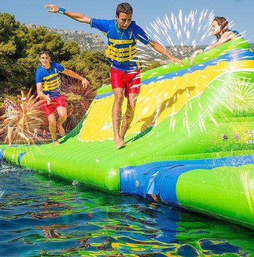 Two boys in life jackets play on a colorful inflatable water slide, while a girl watches from the top, surrounded by fireworks.