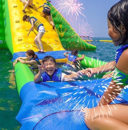 Children enjoying a water play area with inflatable slides, laughing and splashing in the ocean under a sunny sky.