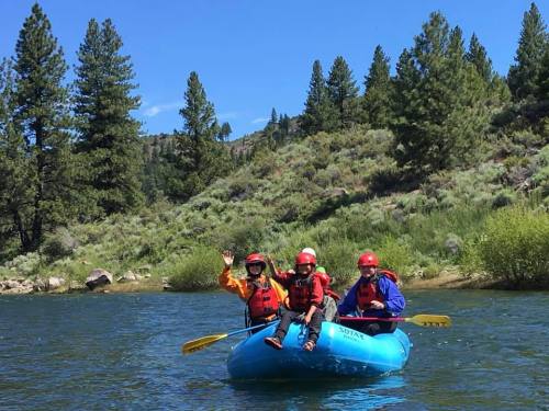 Three people in a blue raft on a river, surrounded by green trees and hills, enjoying a sunny day outdoors.