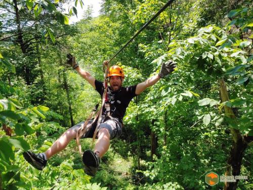 A person in a helmet and gloves zips down a zip line through lush green trees, arms outstretched in excitement.