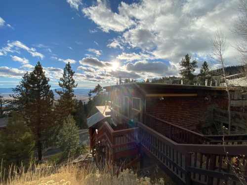 A cabin on a hillside with a view of the valley, surrounded by trees and illuminated by a bright sun in the sky.