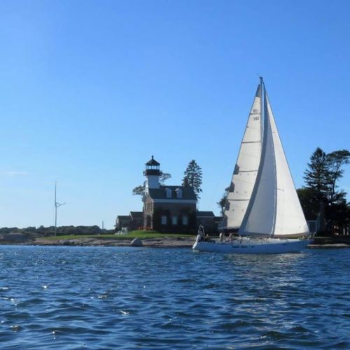 A sailboat glides past a lighthouse on a sunny day, with clear blue skies and calm waters.