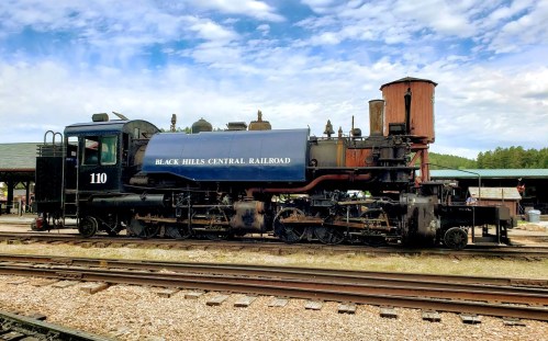 A vintage steam locomotive labeled "Black Hills Central Railroad" parked on tracks with a cloudy sky in the background.