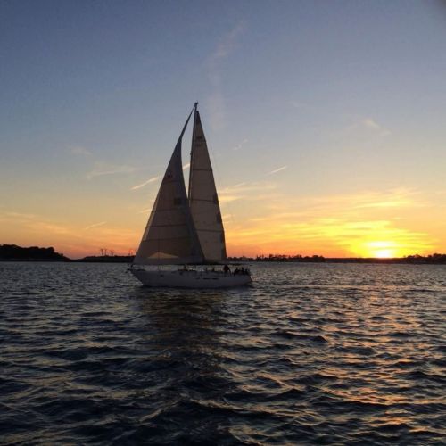 A sailboat glides across the water at sunset, with vibrant orange and purple hues in the sky.