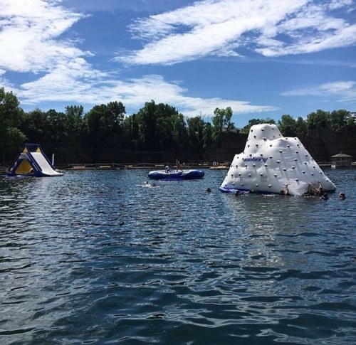 A sunny lake scene with inflatable water structures, including a climbing wall and a slide, and people swimming nearby.