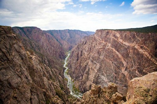 A dramatic view of a deep canyon with steep rock walls and a winding river below, under a partly cloudy sky.