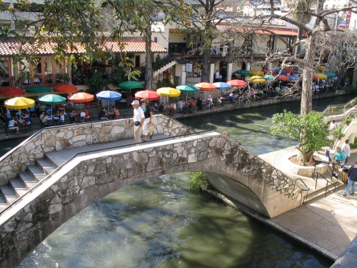 A scenic view of a stone bridge over a river, with colorful umbrellas and people dining along the waterfront.