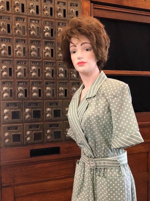 A mannequin in a polka dot dress stands in front of a vintage mail slot wall.