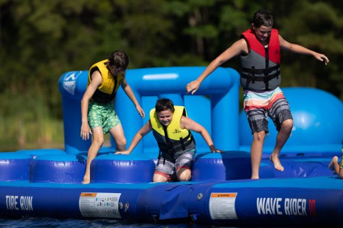 Three kids in life jackets jumping on a blue inflatable water structure at a lake on a sunny day.