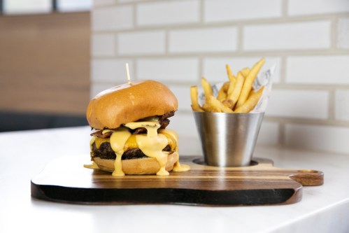 A juicy burger with melted cheese and bacon, served on a wooden board alongside a cup of crispy French fries.