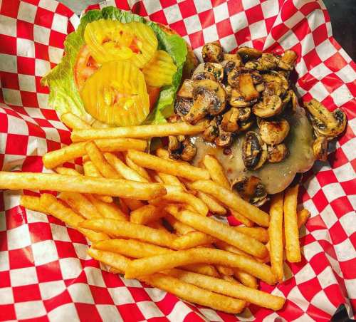 A plate with a burger topped with mushrooms, lettuce, tomato, pickles, and a side of golden French fries in a red checkered basket.