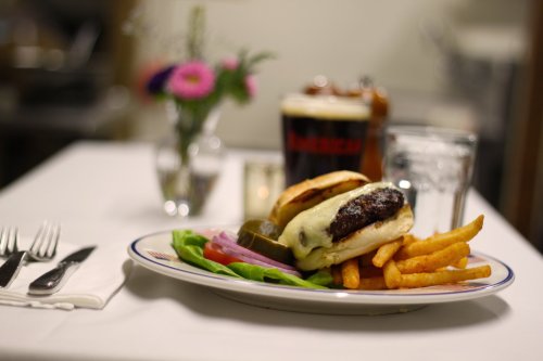 A plate with a cheeseburger, fries, pickles, and onion, with a glass of water and a beer in the background.