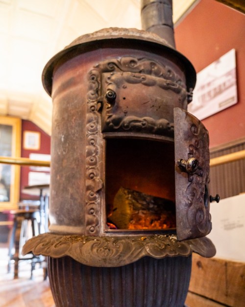 A vintage metal stove with an ornate door, showing glowing embers inside, set against a warm, rustic interior.