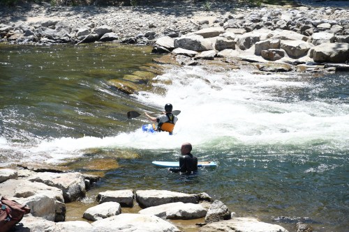 A person in a kayak navigates a small rapid while another person waits on a surfboard in a rocky river.
