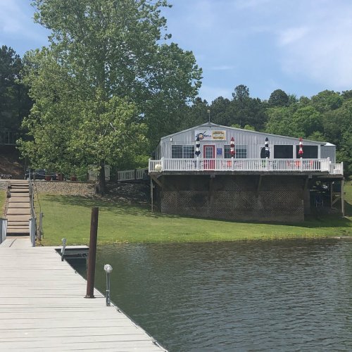 A lakeside view of a building with a deck, surrounded by trees and a wooden dock extending into the water.