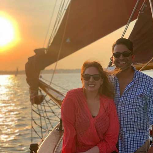 A man and woman smile on a sailboat at sunset, wearing sunglasses, with water and a glowing sun in the background.