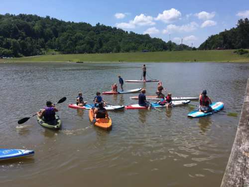 A group of people paddleboarding and kayaking on a calm lake under a sunny sky with green hills in the background.