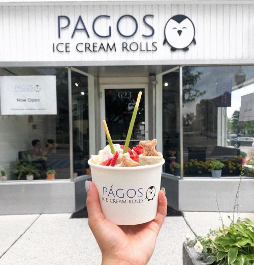 A hand holds a cup of ice cream rolls topped with fruit and cookies in front of the Pagos Ice Cream Rolls shop.