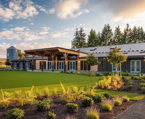 A modern building with a wooden porch, surrounded by lush greenery and trees under a blue sky with clouds.
