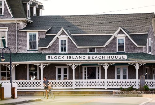A cyclist rides past the Block Island Beach House, a charming gray-shingled building with a white porch.