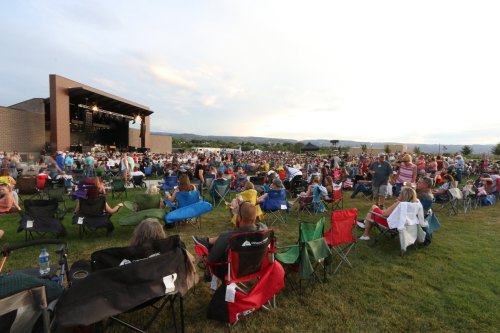 A large crowd enjoys an outdoor concert, seated in lawn chairs on a grassy field with a stage in the background.