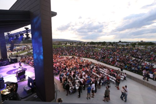 A large crowd gathers at an outdoor concert venue, with a stage and colorful lighting under a cloudy sky.