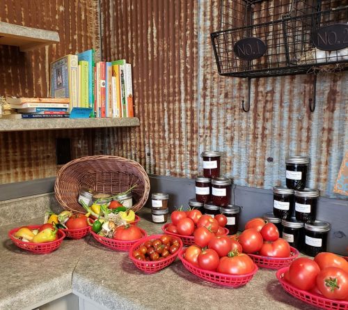 A rustic kitchen counter with baskets of fresh tomatoes, jars of preserves, and a stack of colorful books.