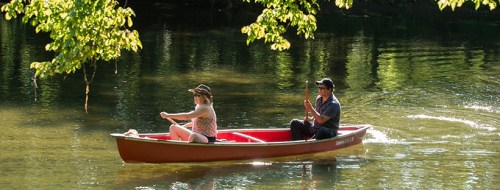 A man and woman paddle a red canoe on a calm river surrounded by green trees and sunlight.