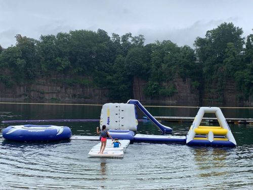 Children playing on an inflatable water park with slides and a floating platform in a calm lake.