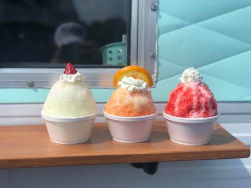 Three colorful snow cones in white cups, topped with whipped cream and fruit slices, displayed on a wooden surface.