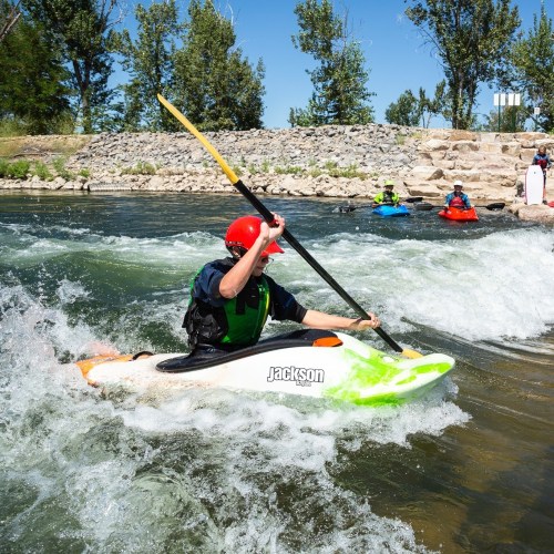 A kayaker paddles through rapids, with other kayakers in the background on a sunny day. Trees line the riverbank.