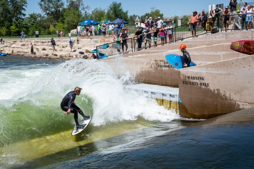 A surfer rides a wave in a river while spectators watch from the bank, enjoying a sunny day.