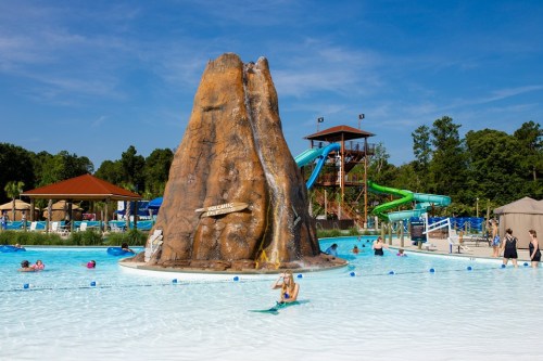 A water park scene featuring a large rock formation, water slides, and people enjoying the pool on a sunny day.